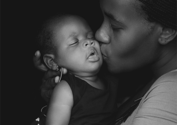 A grayscale photo of a young mother kissing her sleeping baby on the cheek. The baby rests sweetly on the mother.